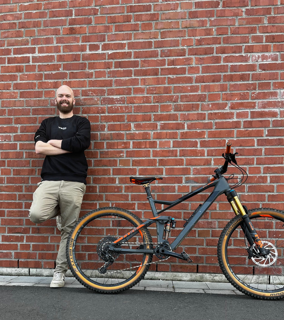 Mann mit velio T-Shirt lehnt sich an einer Ziegelsteinwand neben einem Fahrrad
